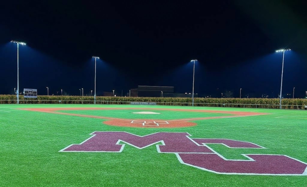 Manheim Central’s baseball field under the lights, edited with corn beyond the outfield fence, and the maroon MC visible behind home plate.